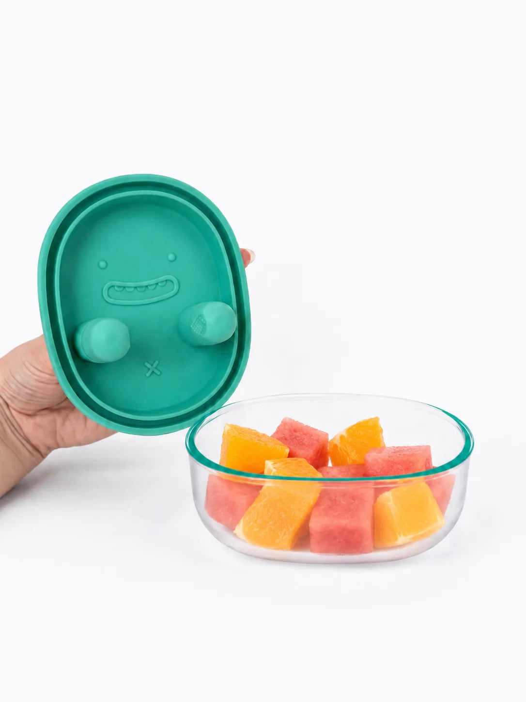 Drobee Snack Miska v pastelově zelené — ruka drží silikonové víčko nad průhlednou miskou s kousky melounu a pomeranče